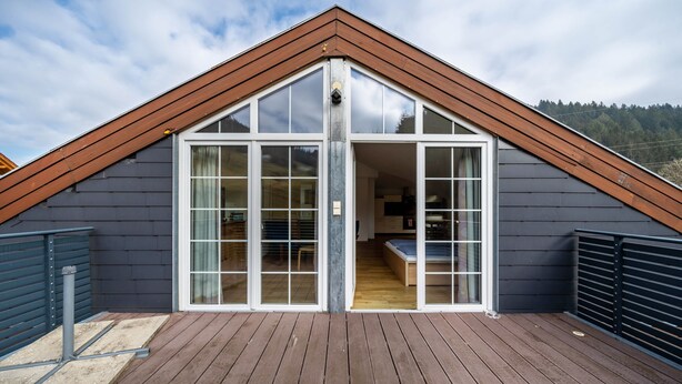 A modern house with a sloped roof, large glass doors leading to a wooden interior, and a deck area. - Holiday Apartment 'Bergblick' with Balcony (Wolfach-Kirnbach)