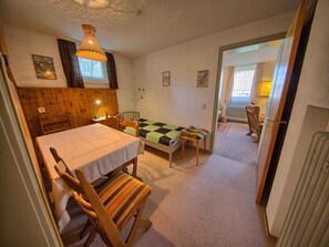 Une pièce avec un lit, une table entourée de chaises, un luminaire suspendu et une vue sur l’extérieur par une porte ouverte.