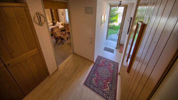 A hallway with a wooden floor, a patterned rug, and a door leading to a bedroom with a bed and chairs. - Apartment 'Kaltenbach' with Shared Terrace, Shared Garden and Wi-Fi (Schonach)