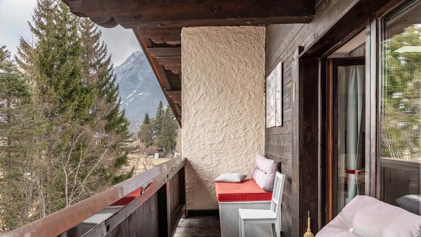 Un balcón con barandilla de madera, un banco acolchado, una silla y un amplio ventanal con vistas a la montaña.
