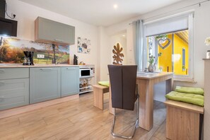 A modern kitchen with grey cabinets, a microwave, and a coffee maker. There is a wooden table with a chair and a window with a yellow wall and a heart decoration. - Holiday Home 'Hedrich' with Private Terrace, Private Garden and Wi-Fi (Gülzow)