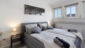 Un dormitorio con cama, mesitas de noche, ventana y un cuadro de un paisaje urbano.