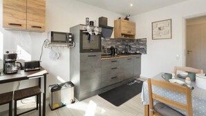 Una cocina compacta con microondas, tostadora y cafetera, una zona de comedor con mesa y sillas, y un cubo de basura pequeño.