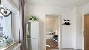 Un pasillo con un radiador blanco, un aparador de vidrio con una planta, una puerta decorada con un corazón y un perchero con una cama al fondo.