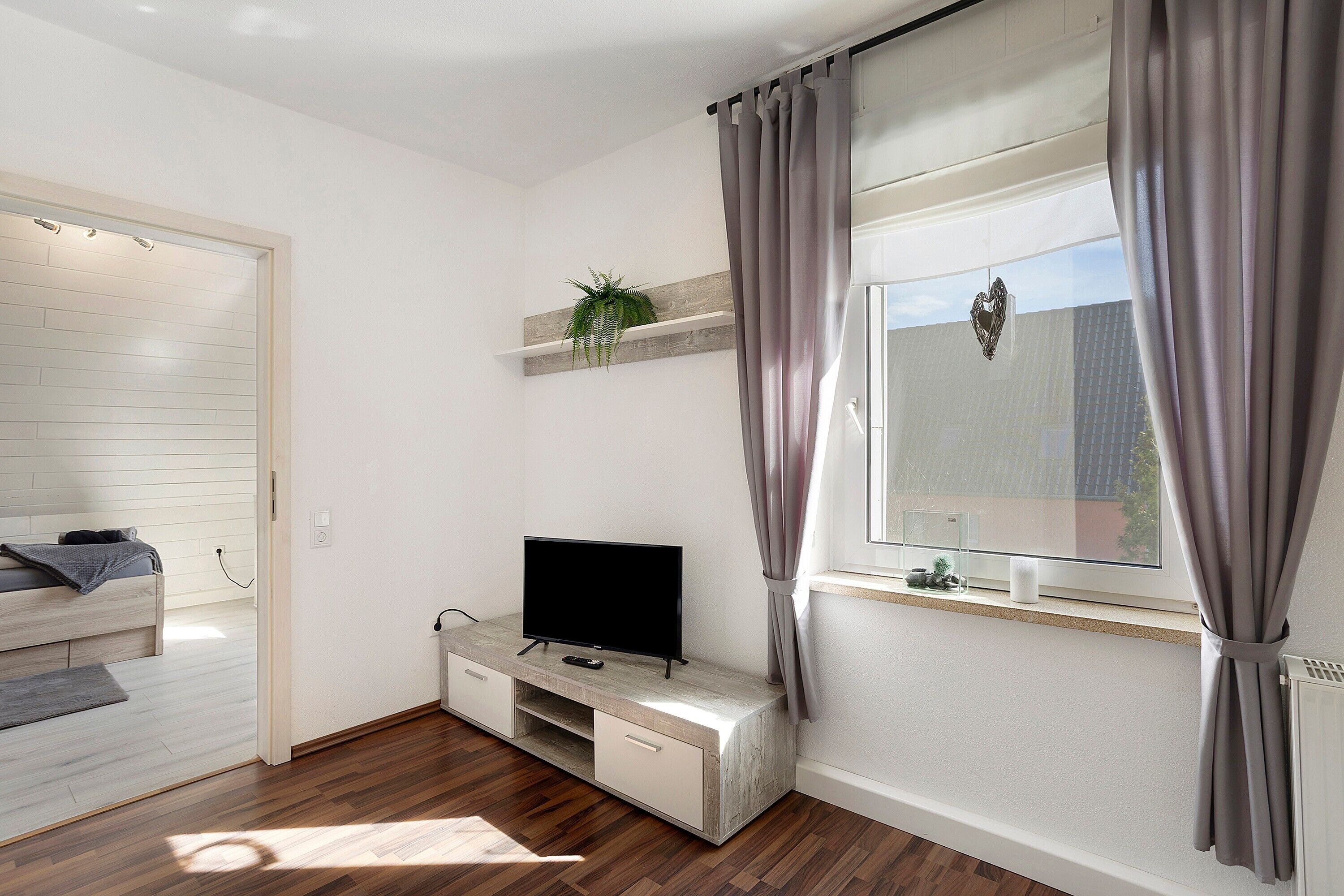 Ein modernes Zimmer mit einem Bett, einem Fernseher auf einer Holzkonsole, einem Fenster mit Vorhängen und einer Pflanze auf dem Regal.