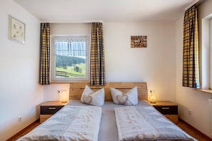 A bedroom with two beds, a window with curtains, a bedside table with a lamp, and a wall-mounted artwork.