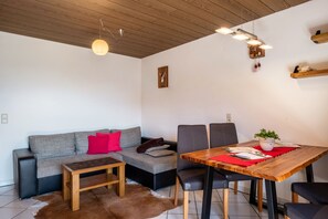 A modern living space with a sofa, a dining table, chairs, a wooden ceiling, and a wall-mounted clock.