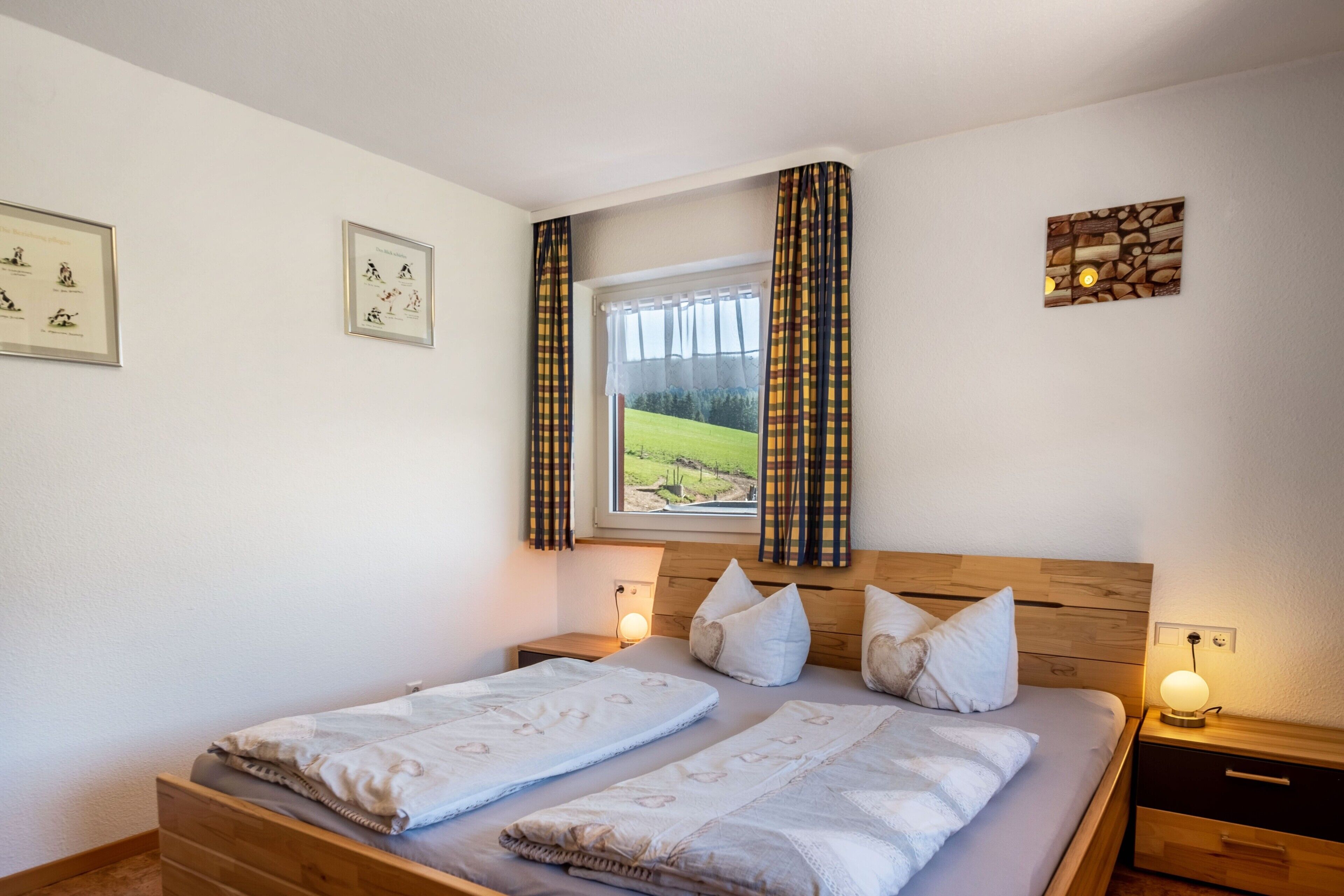 A double bed with a wooden headboard, two bedside tables, a window with curtains, and framed pictures on the wall.