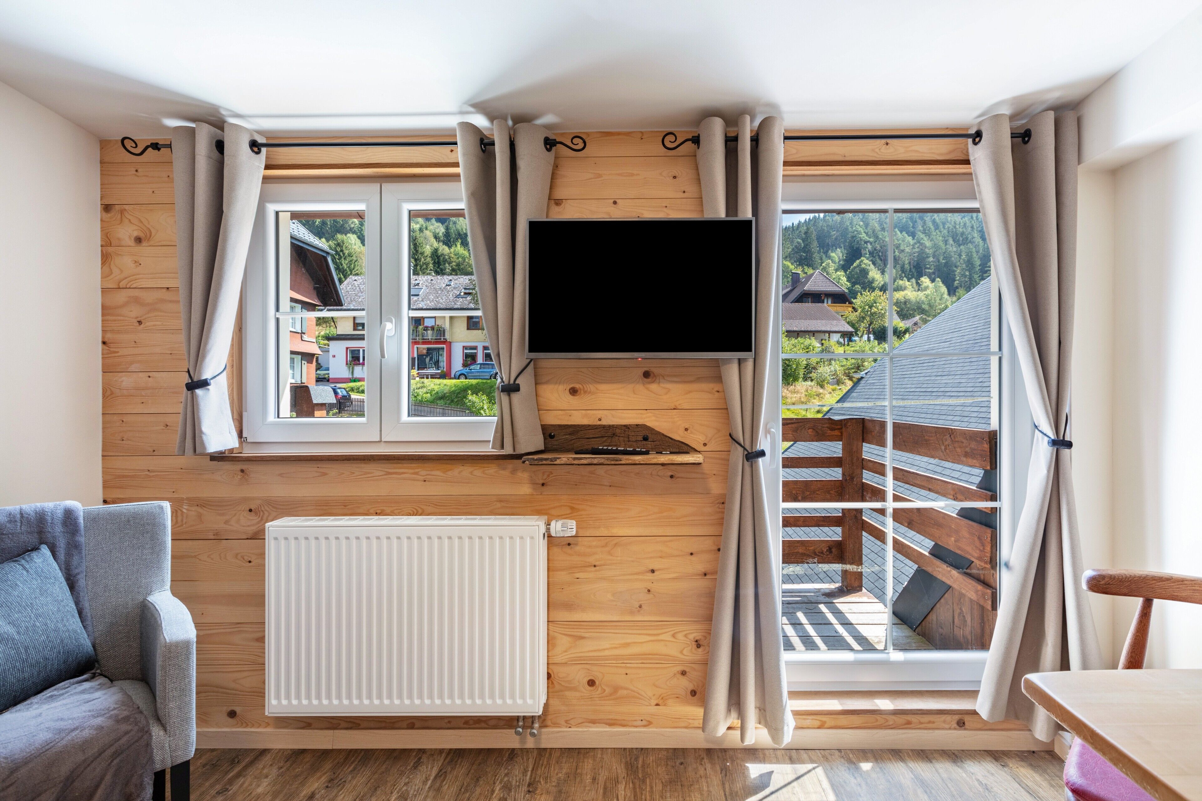 Une pièce avec un mur en bois, un téléviseur fixé au-dessus d’une fenêtre, un radiateur, un canapé et une table.