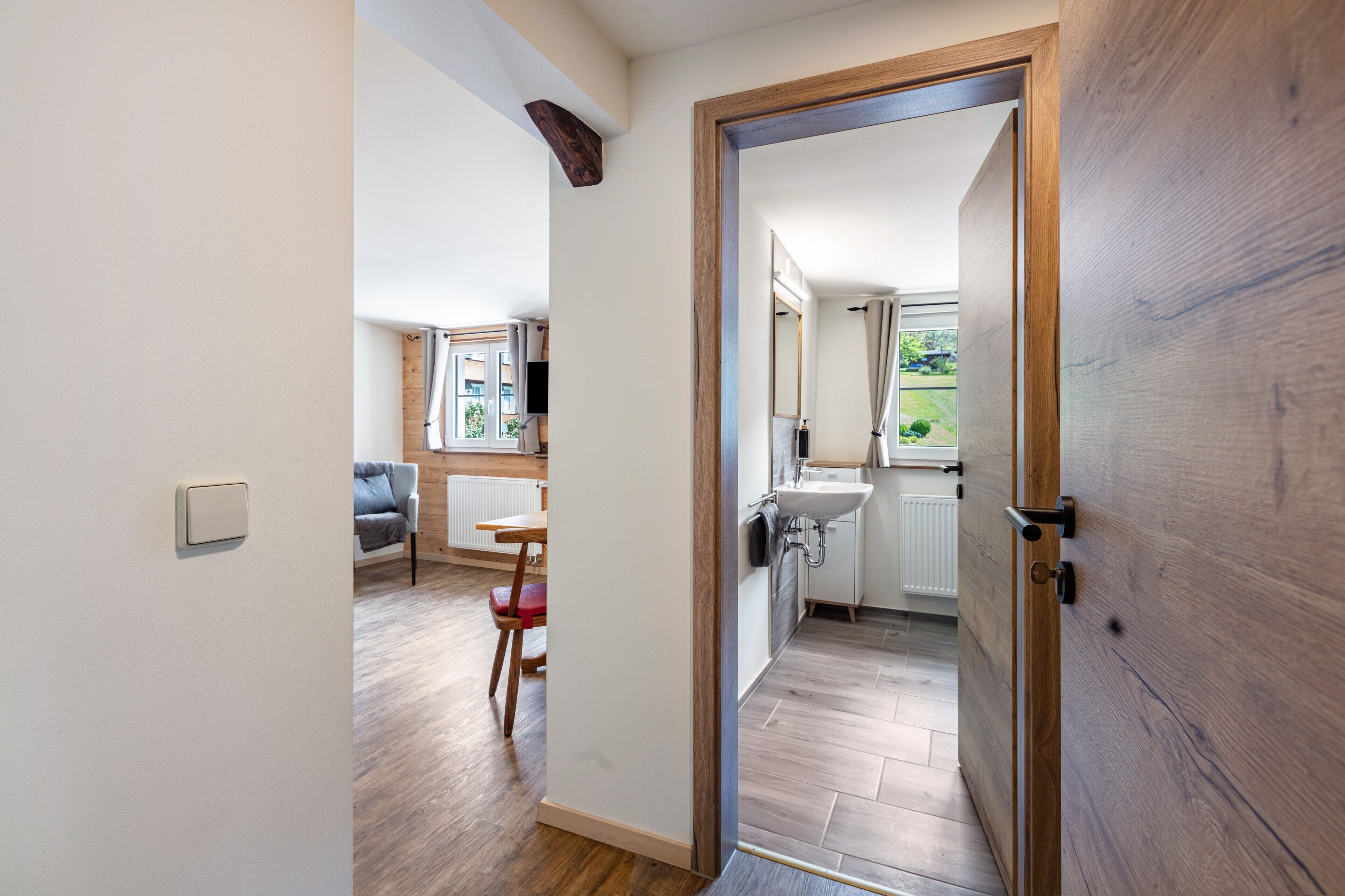 A hallway leading to a kitchen with a wooden door, a dining area with a table and chairs, and a view of the outdoors.