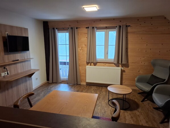 A room with a television mounted on the wall, a radiator, a wooden floor, a round table, and two chairs.