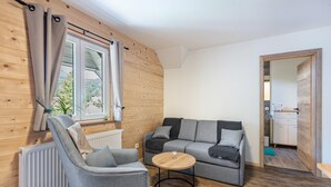 Een moderne woonkamer met een grijze bank, een grijze fauteuil, een ronde houten salontafel en een raam met gordijnen.