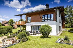 Une maison moderne à deux étages, dotée d’un patio couvert, d’une petite terrasse et d’un jardin bien entretenu.
