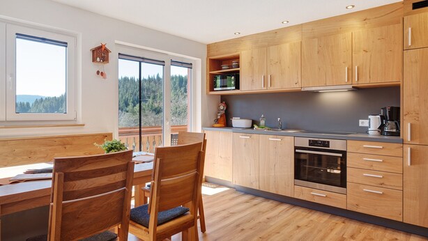 Eine moderne Küche mit Holzausstattung, ein Essbereich mit Tisch und Stühlen sowie ein Blick auf einen Wald durch große Fenster.