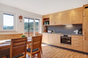 Una cocina moderna con armarios de madera, una zona de comedor con mesa y sillas, y vistas a un bosque a través de grandes ventanales.