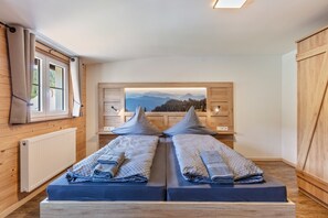 A room with two beds, a wooden floor, a window with curtains, and a view of mountains.