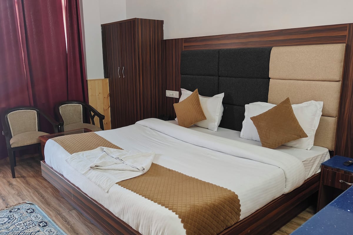 A hotel room with a large bed, two chairs, a wardrobe, and a desk with a remote control.