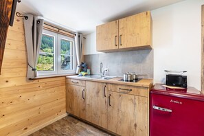 Een compacte keuken met houten kastjes, een spoelbak, een rode Amica koelkast en een raam met gordijnen.