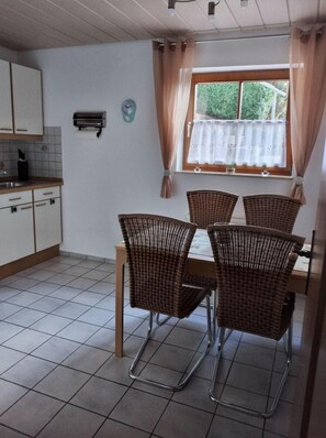 A dining area with a table and four wicker chairs, a kitchen with white cabinets, a clock, and a window with curtains. - Apartment 'Mayntz' Albjuwel' with Shared Terrace, Shared Garden and Wi-Fi (Meßstetten)