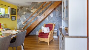 Un coin repas avec une table dressée pour deux personnes, une chaise en osier avec un coussin, un mur en pierre et un escalier en bois.