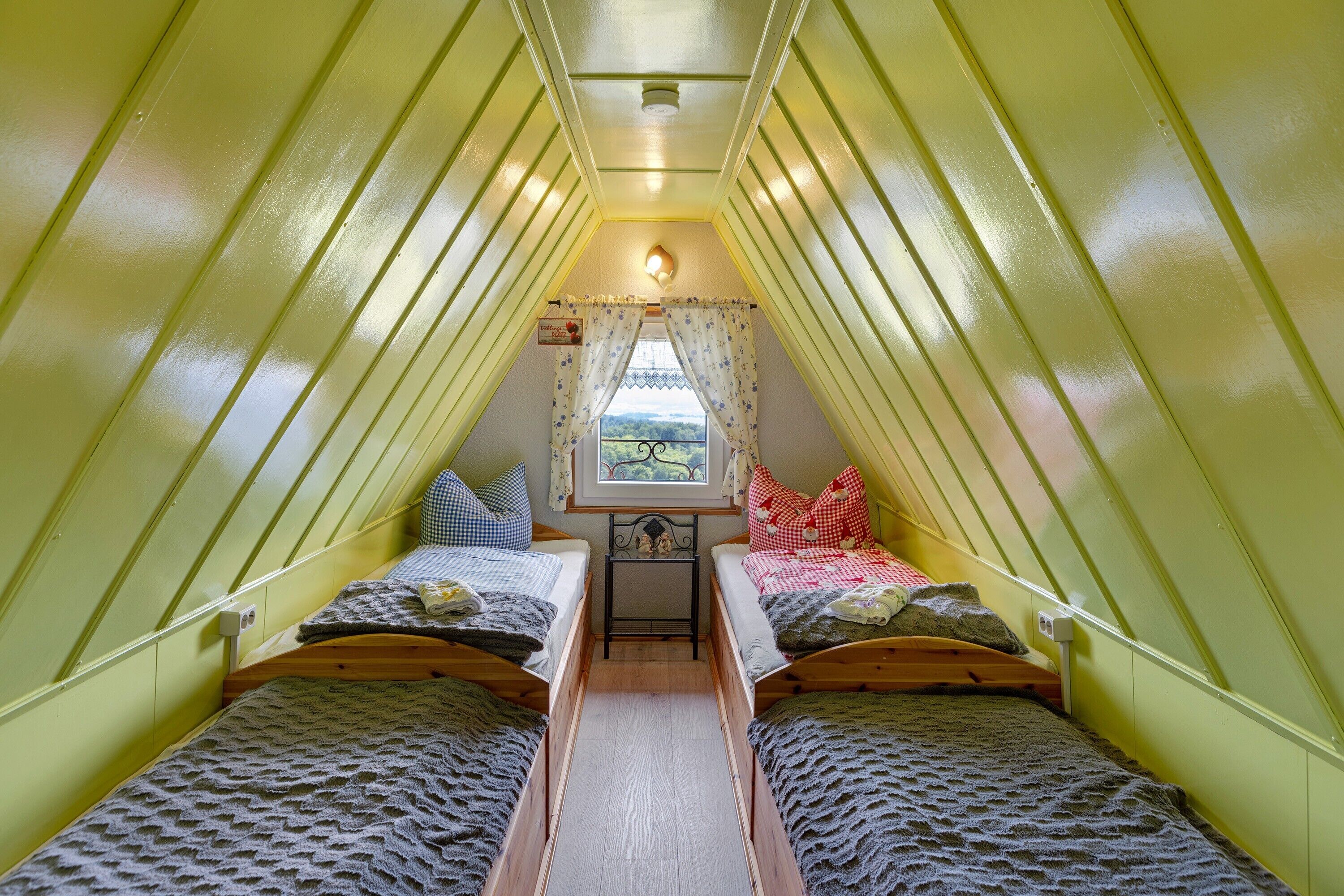 Um quarto de sótão pequeno e triangular, com duas camas, uma janela com cortinas e uma pequena mesa.