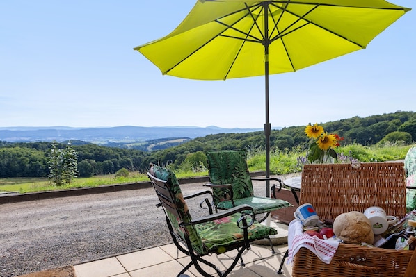 A wicker chair and table set under a yellow umbrella, with a wicker basket containing food items and a vase of sunflowers.