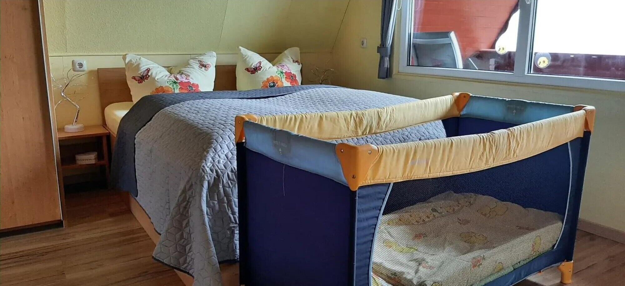 Ein Zimmer mit zwei Betten, einem Fenster, Holzboden und einem kleinen Tisch mit Lampe.