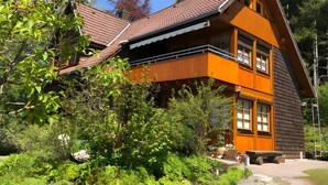 Ein zweistöckiges Holzhaus mit geneigtem Dach, umgeben von üppigem Grün.