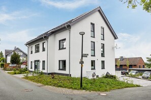 Un immeuble résidentiel moderne à deux étages, doté d’une façade blanche, de nombreuses fenêtres et d’un petit jardin agrémenté de verdure.