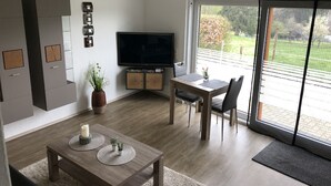 Una sala moderna con televisor de pantalla plana, una mesa de comedor con sillas, una planta en maceta y un amplio ventanal corredero que da a un balcón.