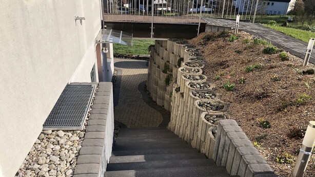 Una escalera de concreto con una reja de metal a la izquierda, que conduce a un área pavimentada con un borde de piedra y una pendiente cubierta de césped y plantas.