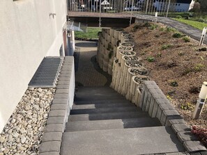 A concrete staircase with a metal grate on the left, leading to a paved area with a stone border and a grassy slope with plants.