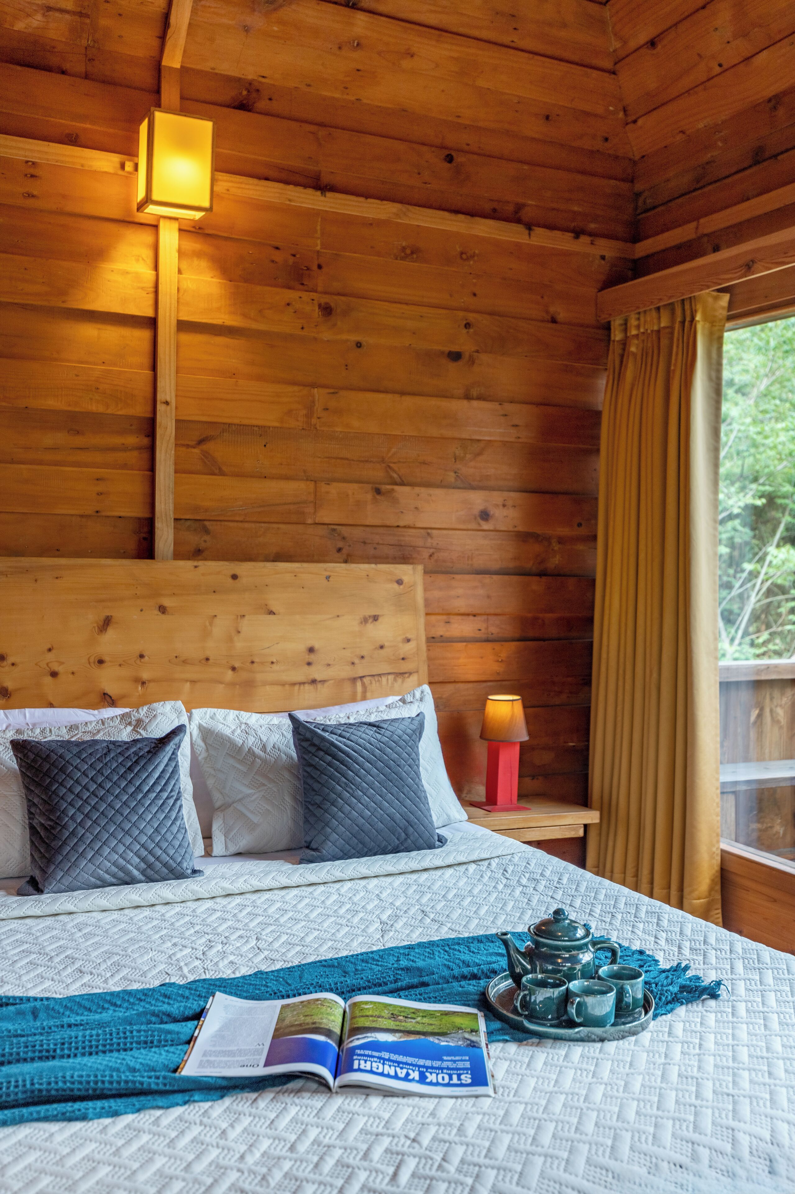 A wooden cabin bedroom with a bed, pillows, a reading light, a window with curtains, and a tea set on the bed.