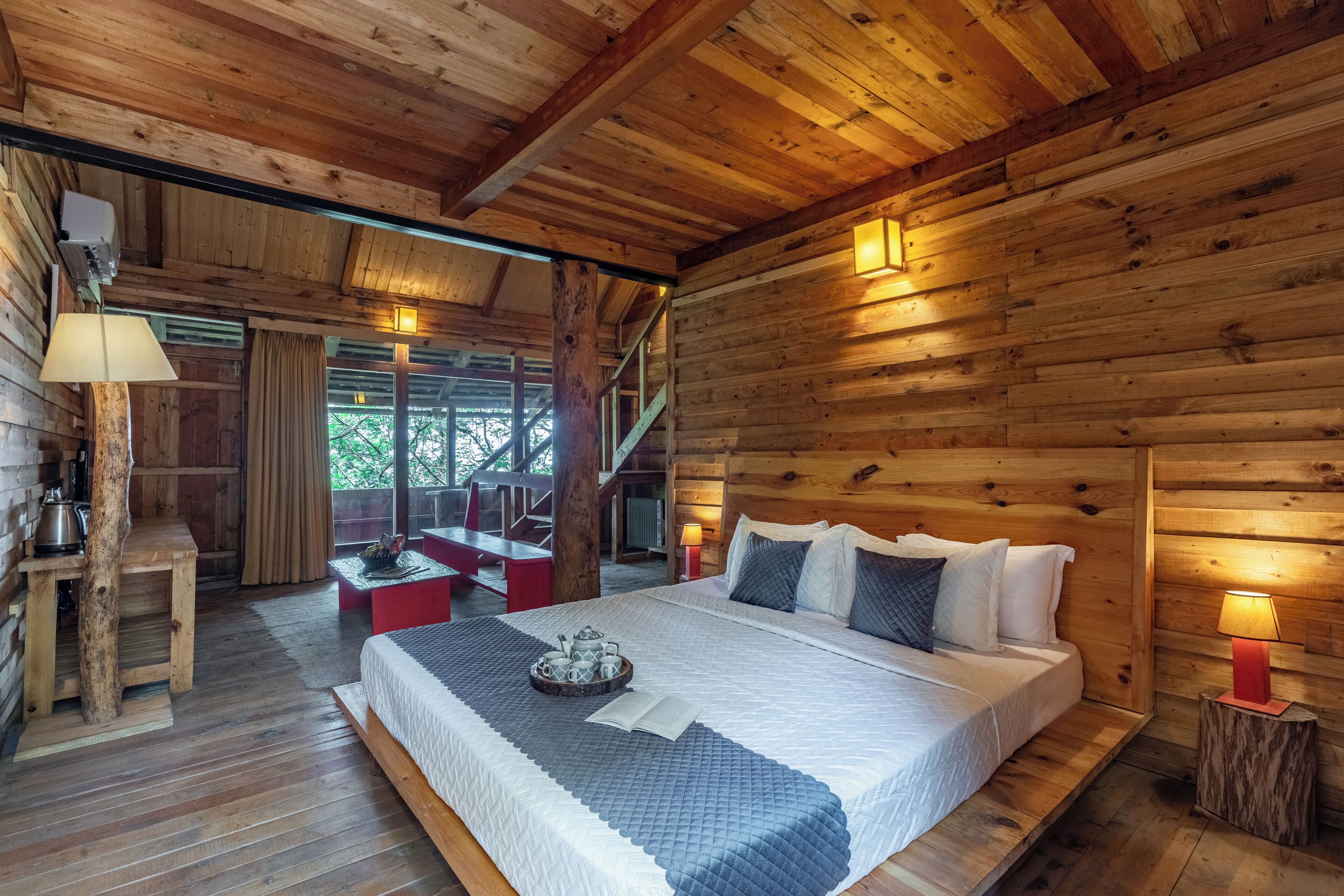 A wooden cabin bedroom with a large bed, a small table with a tray of tea, and a staircase leading to an upper level.