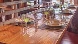 A wooden dining table set with glasses, plates, and cutlery, with a stone wall and wooden beams in the background. - Alaya Stays Suro Woodcrest Chalet (Kandaghat)