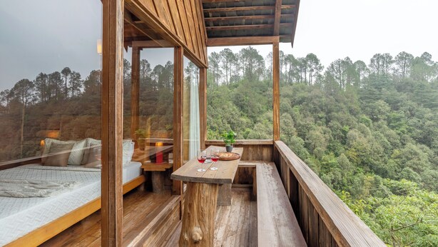 A wooden balcony with a bed and a table, overlooking a forested hillside. - Alaya Stays Suro Woodcrest Chalet (Kandaghat)