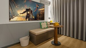 Ein modernes Hotelzimmer mit einem großen Poster eines Videospielcharakters, einer Holzbank, einem kleinen runden Tisch und einem Mülleimer.