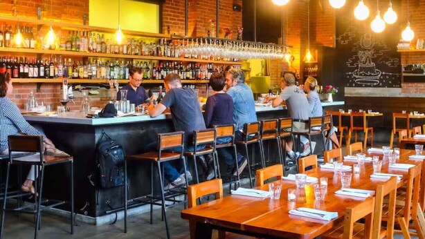 Eine Barbereich mit Backsteinwand, hängenden Glühbirnen und einer Bar mit Flaschen und Gläsern.