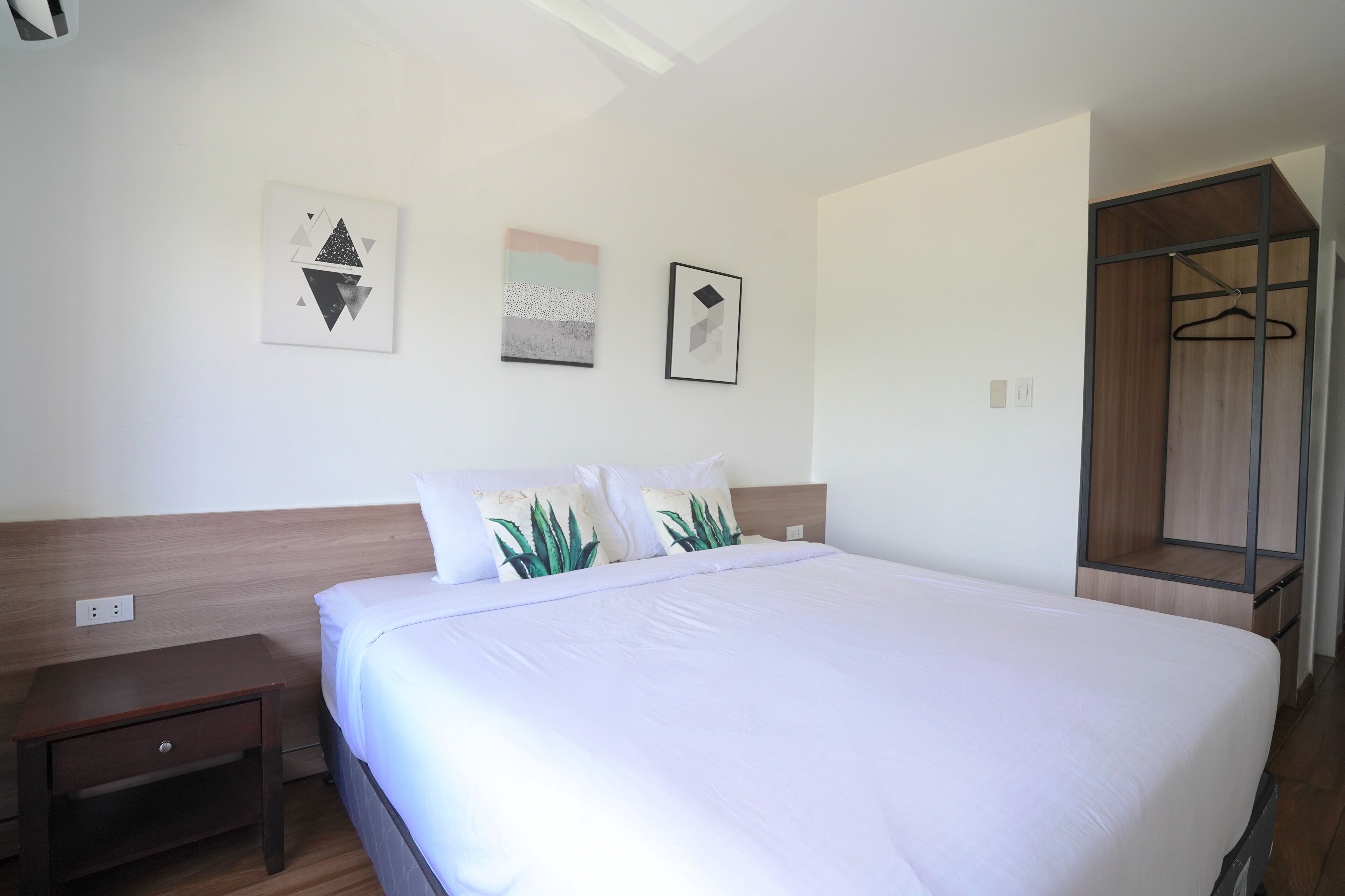 A neatly made bed with white bedding, a wooden headboard, and two pillows with plant designs. There are three framed artworks on the wall above the bed.