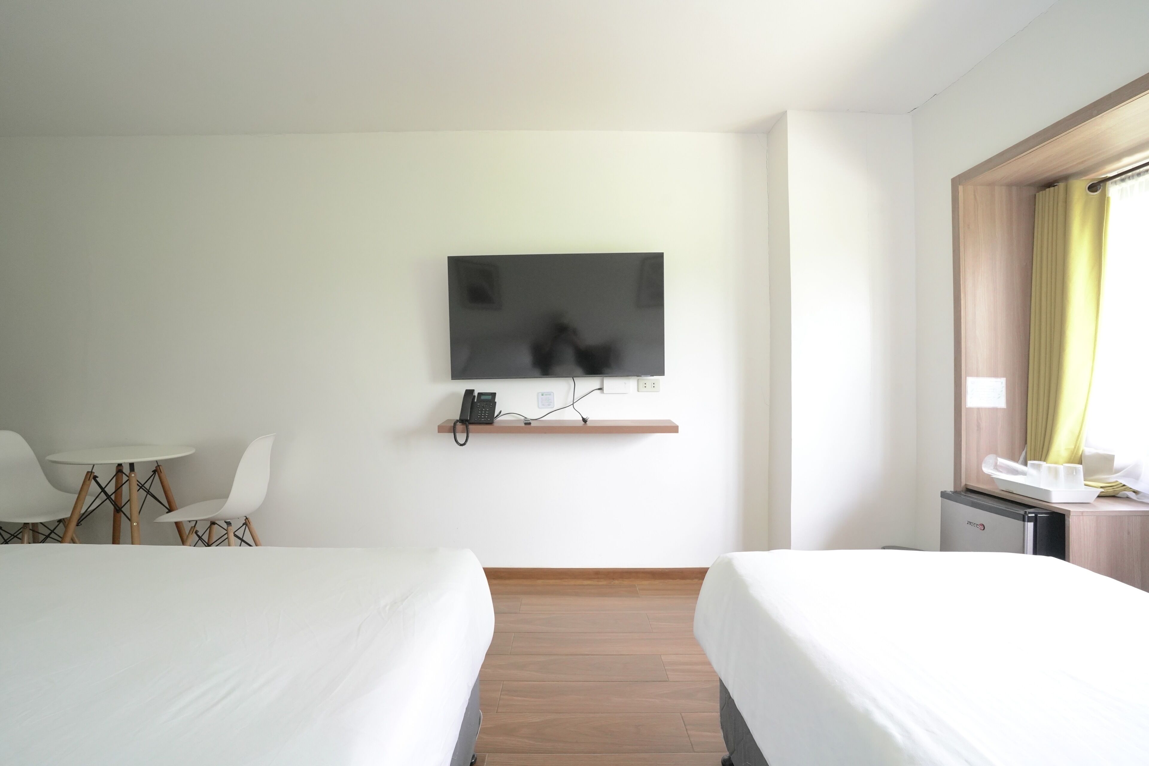 A hotel room with two beds, a TV mounted on the wall, a small desk with a phone, and a window with curtains.