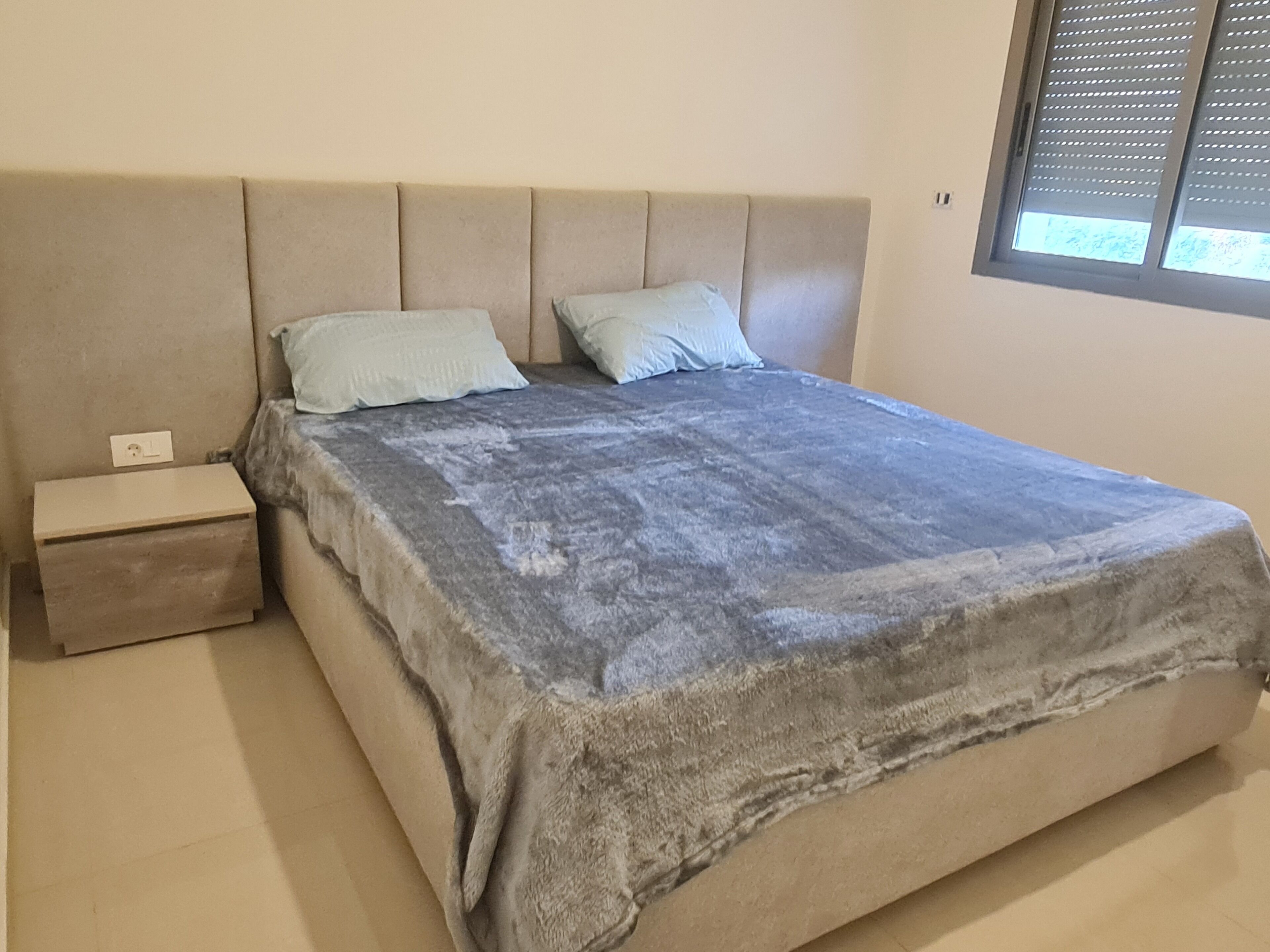 Een slaapkamer met een bed, twee kussens, een nachtkastje en een raam met jaloezieën.