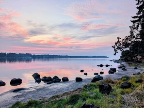 A lakeside view with a calm water body, a rocky shoreline, and a distant tree line.