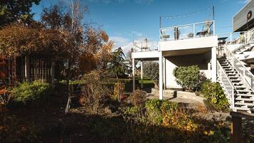 Un espace extérieur abrité, doté d’un escalier menant à un balcon situé au deuxième étage, et entouré de végétation et d’arbres.