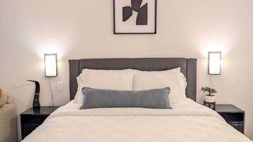 Ein ordentlich bezogenes Bett mit einer weißen Decke und einem einzelnen blauen Kissen, begleitet von zwei Nachttischen mit Lampen, auf einem steht eine Pflanze.