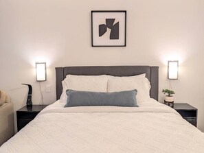 A neatly made bed with a white quilt and a single blue pillow, flanked by two bedside tables with lamps, one of which has a plant on it.