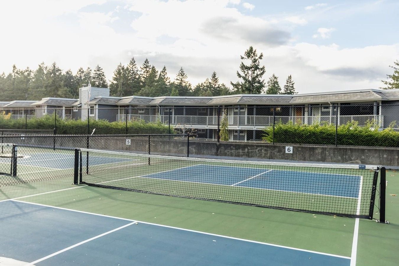 A tennis court with a net, surrounded by a fence, and adjacent buildings with balconies.