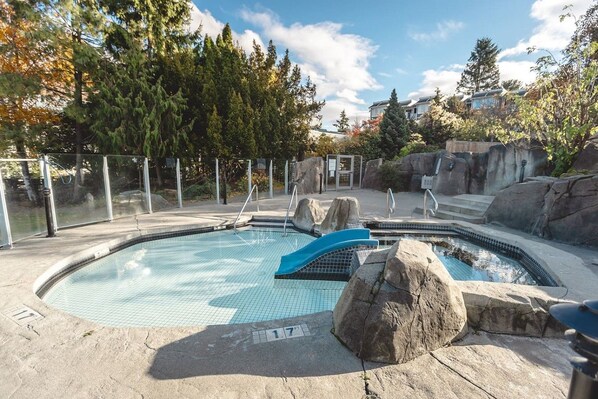 A swimming pool with a rock feature, surrounded by a fence, and trees in the background.