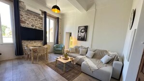 Uma sala de estar com sofá, mesa de centro, mesa de jantar com cadeiras, parede de pedra, janela com cortinas e luminária pendente.