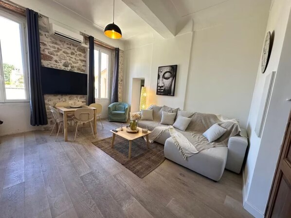 A living room with a sofa, a coffee table, a dining table with chairs, a stone wall, a window with curtains, and a hanging pendant light.