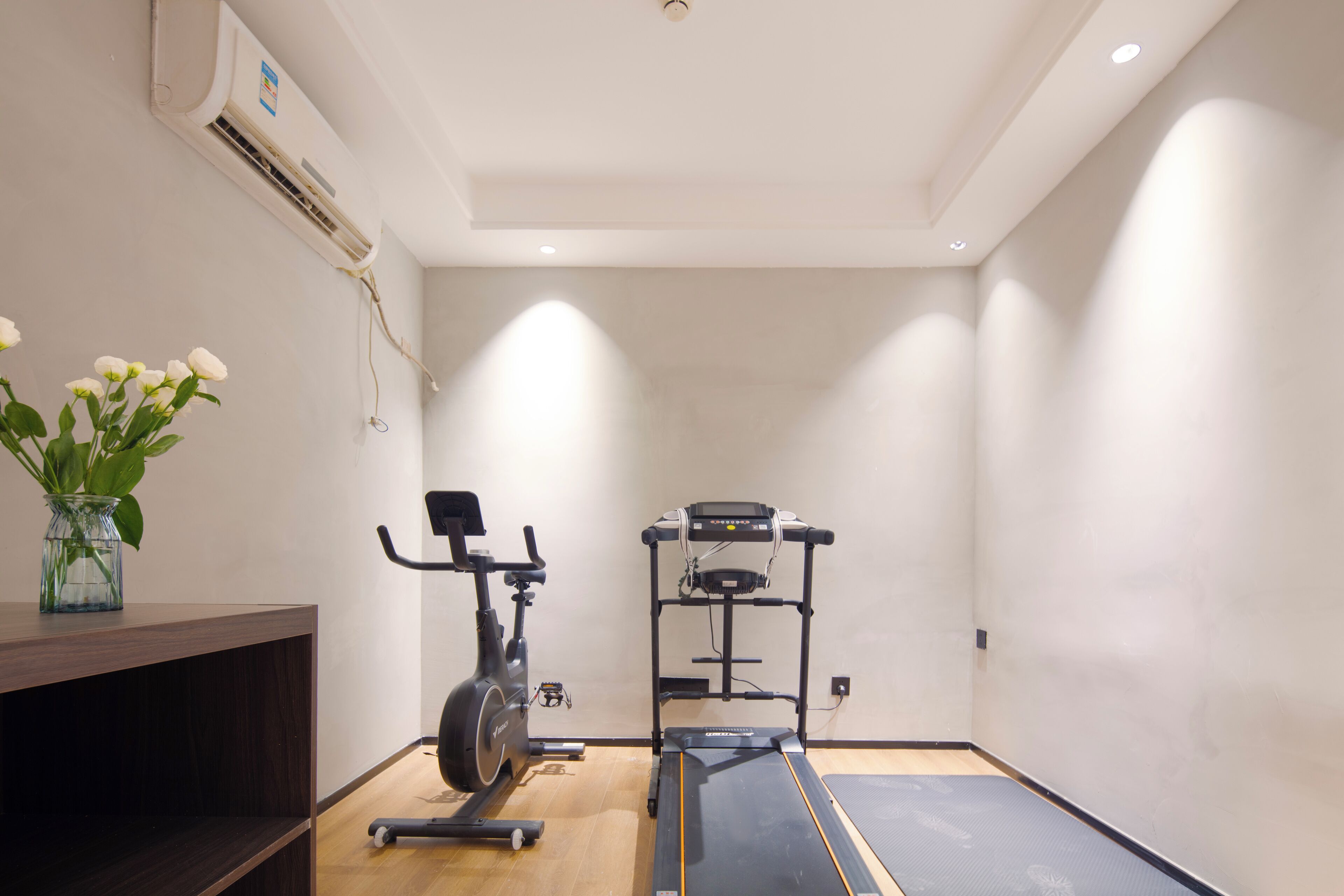 A small, well-lit room with a treadmill, a stationary bike, and a wall-mounted air conditioning unit.
